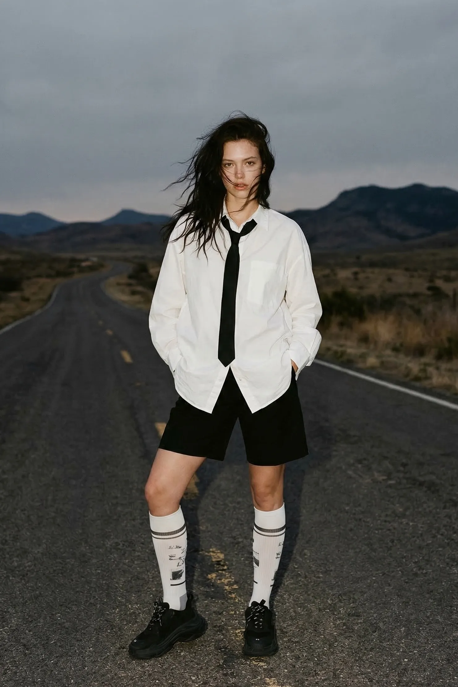 A full-body shot captures a young woman with light skin and long, dark, tousled hair, standing confidently on a deserted…